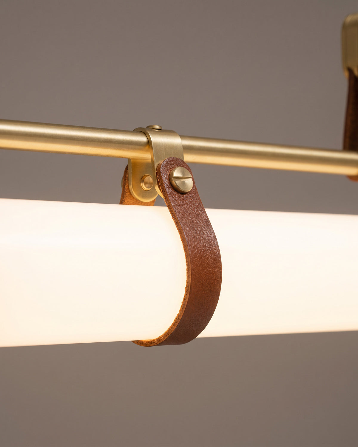 Suspension Boucle: gros plan sur une sangle brun cognac enroulée autour du tube opalin sous la barre dorée